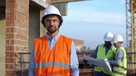 Portrait-of-the-handsome-Caucasian-man-in-hardhat-and-goggles-smiling-and-posing-to-the-camera-at-the-building-site.-Two-multiethnic-colleagues,-man-and-woman-talking-about-work-on-the-background.